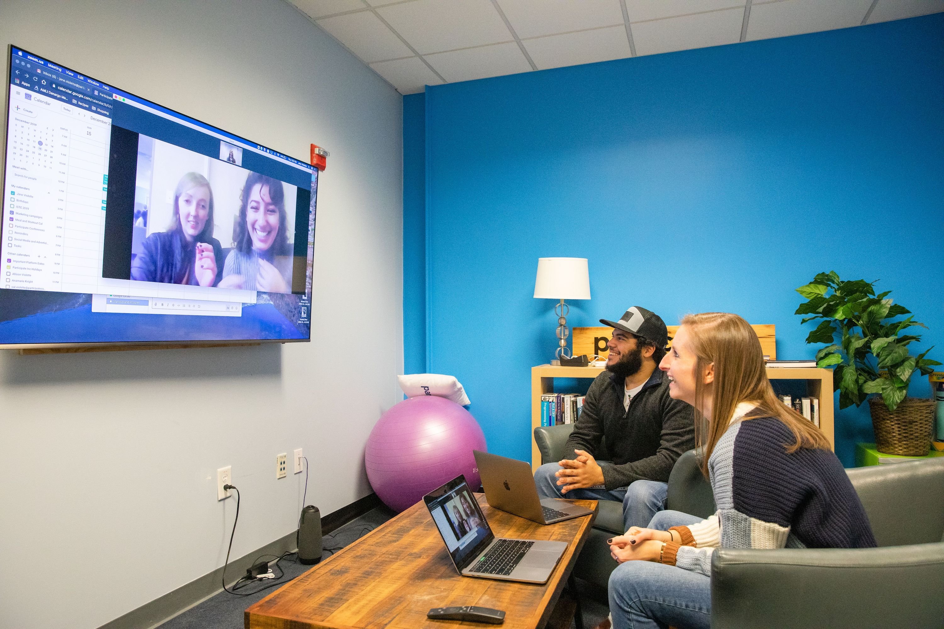 Two team members sitting in chairs smiling at a TV screen, where two other team members are shown. They are all on a video call.
